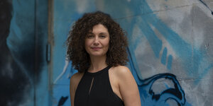 Die Violinistin Biliana Voutchkova steht vor einer Wand mit abstrakten Graffiti in verschiedenen Blautönen, sie trägt ein schwarzes Kleid