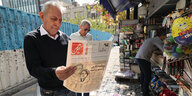 Ein Mann steht auf einer STraße am Kiosk und hält eine Zeitung, 2 gezeichnete Frauen auf dem Titel