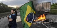 Eine Frau mit einer Brasilienfahne steht auf einer Autobahn, hinter ihr brennt eine Barrikade