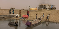 ein Büffel im Wasser, ein Boot daneben, Kinder und Männer stehen auf einem trockenen Land, ein roter Wasserkanister am Ufer