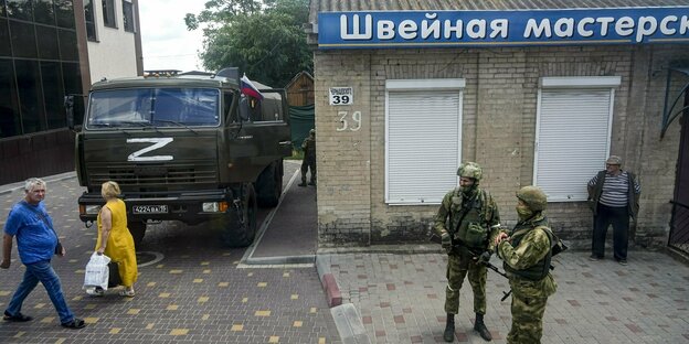 Russische Soldaten und ein russisches Militärfahrzeug vor einem Geschäft, Zivilisten laufen vorbei