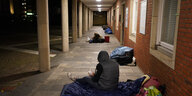 Personen in dicken Jacken sitzen und liegen auf Schlafsäcken im Eingangsbereich eines Gebäudes.