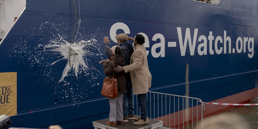 Drei Menschen stehen vor der Sea-Watch 5 und haben eine Sektflasche dagegen geschwungen, die gerade am Rumpf schäumend explodiert.