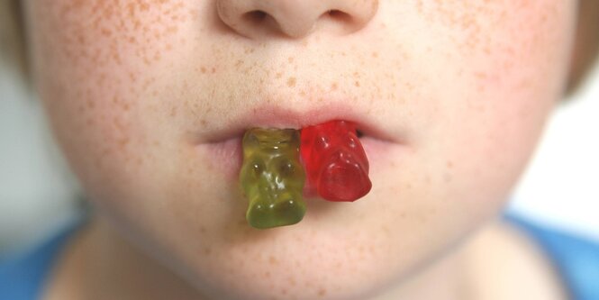 Detailaufnahme eines Kindergesichts. Das Kind hat ein rotes und ein grünes Gummibärchen zwischen den Lippen