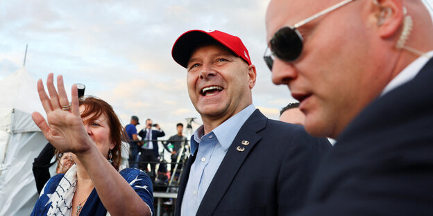 Republican candidate Doug Mastriano in a red flat cap and smiling victoriously.  In the foreground a security officer