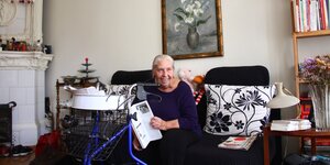 Eine ältere Frau mit langen grauen Haaren auf ihrem Sofa, davor ein Rollator, an der Wand hängt ein Bild