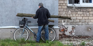 ein Mann lädt zwei kleinere Baumstämme auf sein Fahrrad, das an einer Hauswand lehnt