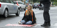 Eine junge Frau sitzt auf der Straße, nebn ihr sind die Beine eines Polizisten zu sehen