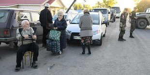 Ein Mann mit Krücken sitzt auf einem Stuhl auf der Straße, eine Frau mit Gepäck schaut verloren, ein Auto mit Z parkt, Soldaten stehen auf der STraße