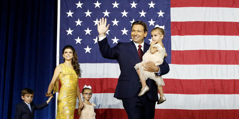 Gouvereur Ron DeSantis betritt mit seiner Familie die Bühne. Er trägt eines der Kinder auf dem Arm, die Frau hat die beiden anderen Kinder an der Hand. Im Hintergrund eine US-Flagge