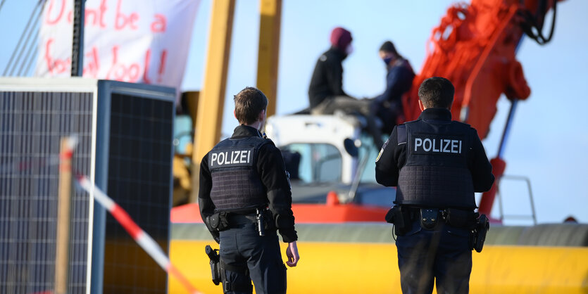 Zwei Polizisten schauen auf die Baustelle. Dort haben Aktivist:innen Transparente aufgehängt und Fahrzeuge besetzt