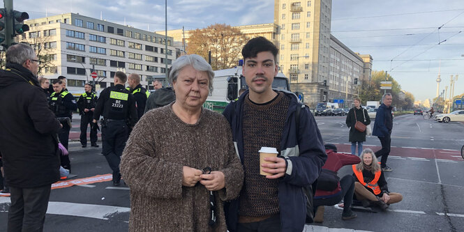 Eine alte gedrungene Frau mit Pullover und Grauen Haaren sieht auf einer Straßenkreuzung neben einem jungen Mann mit Jacke und Kaffeebecher gemeinsam mit ihm in die Kamera. In dem Hintergrund bewegen sich Polizisten vor einem Hochhaus, was noch weiter im Hintergrund steht, und eine Frau mit orangener Weste sitzt auf dem Boden.