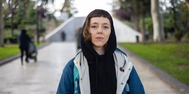 Eine junge Frau mit kurzem Pony und Nasenring trägt ein schwarzes Kapuzenshirt und eine Outdoorjacke