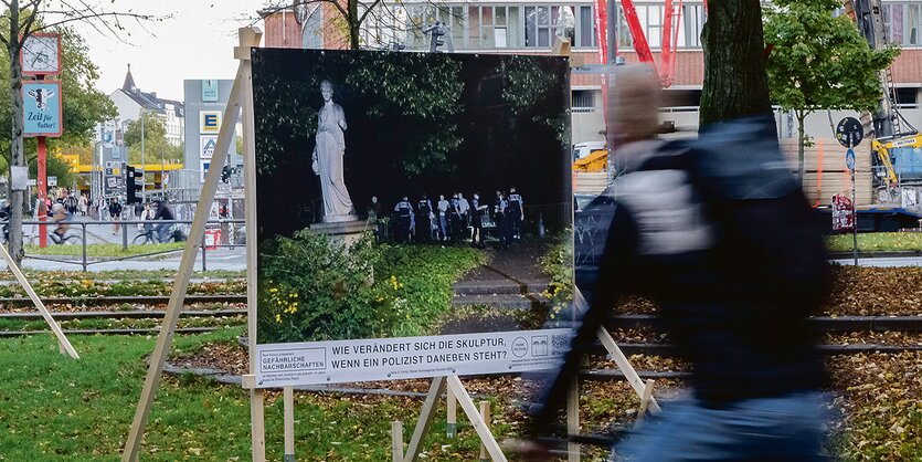 Ein Radfahrer fährt an einem Poster vorbei, auf dem eine Skulptur nachts in einem Park zu sehen ist. Neben der Skulptur stehen Polizisten.