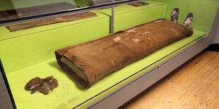 Ein Stück Kopfhaut mit Haaren liegt in einer Vitrine. Daneben ein Umhang und Sandalen