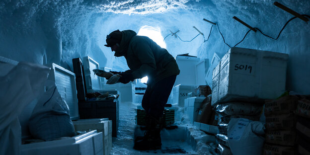 Ein Mann steht gebückt in einer Eishöhle und packt etwas aus einer Kiste aus, viele Kisten einer Froschungsstation