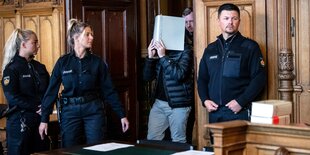 Polizist*innen begleiten den Angeklagten S. in den Gerichtssaal, er hält sich eine Mappe vors Gesicht