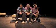 Drei Männer sitzen auf einer Bühne, einer spielt Saxofon