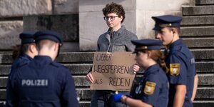 Ein Klimaaktivist der Gruppe "Aufstand der letzten Generation" ist von Polzisten und Polizistinnen umringt. Er hält ein Protestplakat auf dem zu lesen ist "Lieber wegsperren als reden"