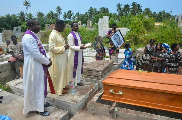 Three priests in robes stand at the grave.  The coffin is laid out in front of them.  Next to the men is a woman in festive clothes with a photo of the deceased.  Next to it another person dressed in festive clothes.  Behind a few men.