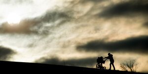 Eine Frau schiebt unter turbuöent wolkigem Himmel einen Kinderwagen einen Hügel hinauf