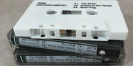 Eine weiße Kassette mit der Aufschrift „Stau / Großraumbüro“ liegt auf zwei Kassettenhüllen, auf deren Rücken verschiedene Titel des Albums wie „A1 The Scam“ zu lesen sind