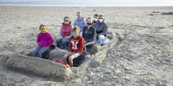 Kinder sitzen in einem aus Sand gebauten Bus am Strand und tragen Masken