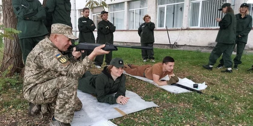 Ein Mann zielt mit einer Waffe, neben ihm liegen lachend und lächelnd zwei junge Frauen auf dem Boden, in Schieß-Position. Vor der einen liegt eine Schusswaffe