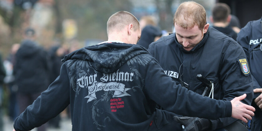 Neonazis mit Thor Steinar-Pulli wird von einem Polizisten kontrolliert