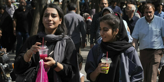 2 Frauen gehen auf der Straße, sie haben das Kopftuch abgelegt, halten einen Getränkebecher ind er Hand