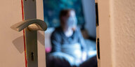 Blick durch die Tür: Eine Frau sitzt während der Isolation nach einem positiven Corona-Test mit Maske auf ihrem Bett