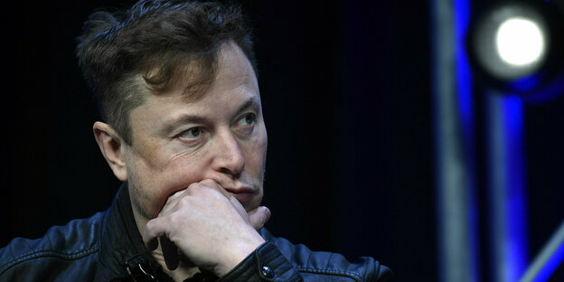 Eine Zeichnung von Elon Musk. Er sitzt auf einem Stuhl, trägt Anzug und Krawatte. Vor ihm ein Mikrofon.