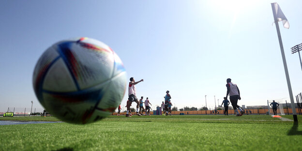 Der WM Ball fliegt über en Platz, im Hintergrund Fussballer die trainieren