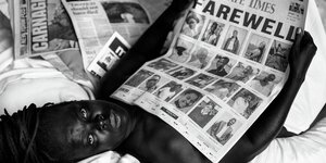 Ein Mensch liegt auf einem Bett, in der Hand eine Zeitungsseite mit vielen Porträts