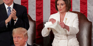 Nancy Pelosi zerreißt einen Stoß Papier, vor ihr steht Donald Trump