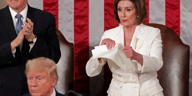 Nancy Pelosi zerreißt einen Stoß Papier, vor ihr steht Donald Trump