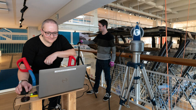 Vermessungsingenieurin Heidi Hastedt steht über einen Laptop gebeugt. Im Hintergrund sind ein wissenschaftlicher Mitarbeiter, Messinstrumente, und die Planken der Bremer Kogge zu sehen.