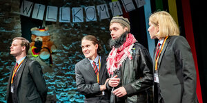 Vier Menschen auf einer Bühne, drei von ihnen tragen Anzüge und Anhänger an Bändern mit Deutschlandfahne. Einer trägt eine Lederjacke, eine Mütze und ein Tuch um den Hals