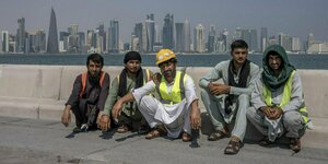 Fünf Arbeiter sitzen auf Betonbänken am Meer vor der Skyline von Doha. dEr Mann in der Mitte trägt einen Bauhlem, die anderen nur Signalwesten