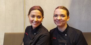 Mona Warnecke und Kelly Alpers sitzen auf einer Bank in ihrer Konditoreifiliale.