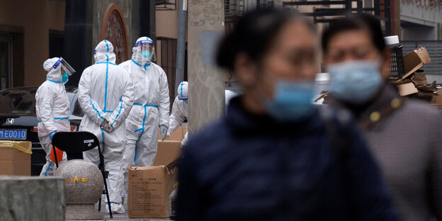 Epidemic personnel in protective suits stand on the street, in the foreground passers-by with masks are photographed out of focus