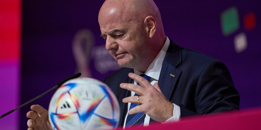 Infantino sitzt vor dem Rednerpult breitet Hände aus, dazwischen liegt ein Fußball