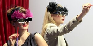 Zwei junge Frauen verkleidet mit Federn und altmodischen Datenbrillen, eine zielt auf etwas