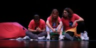 Drei Performer:innen sitzen auf dem Boden einer Bühne