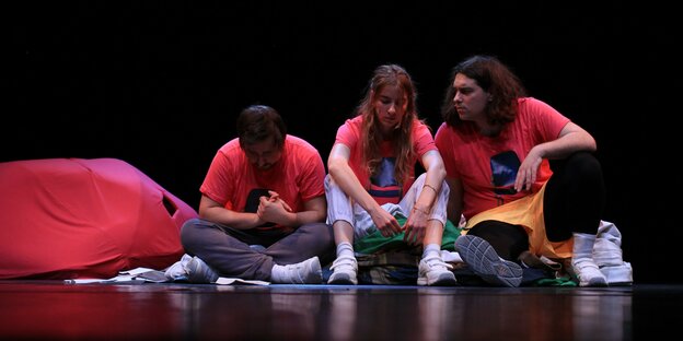 Drei Performer:innen sitzen auf dem Boden einer Bühne