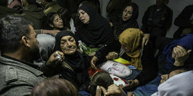 Weinende und trauernde Frauen umringen den Sarg eines Mannes, dessen Foto groß auf dem Sarg liegt