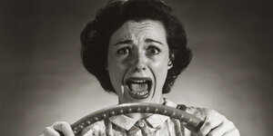 Schwarz-Weiß-Fotografie einer Frau mit aufgerissenem Mund am Steuer eines Autos