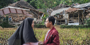 Zwei Personen halten sich an den Händen, im Hintergrund ein zerstörtes Haus.