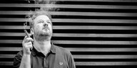 Der Musiker Stefan Bethke steht vor einer vertäfelten Wand, er hält eine Zigarre in der Hand und schaut dem Rauch nach, der nach oben wegzieht