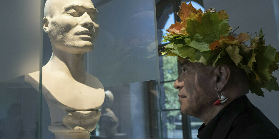 Ein Mann mit einem Kranz aus Blättern schaut auf eine Büste in einer Glasvitrine